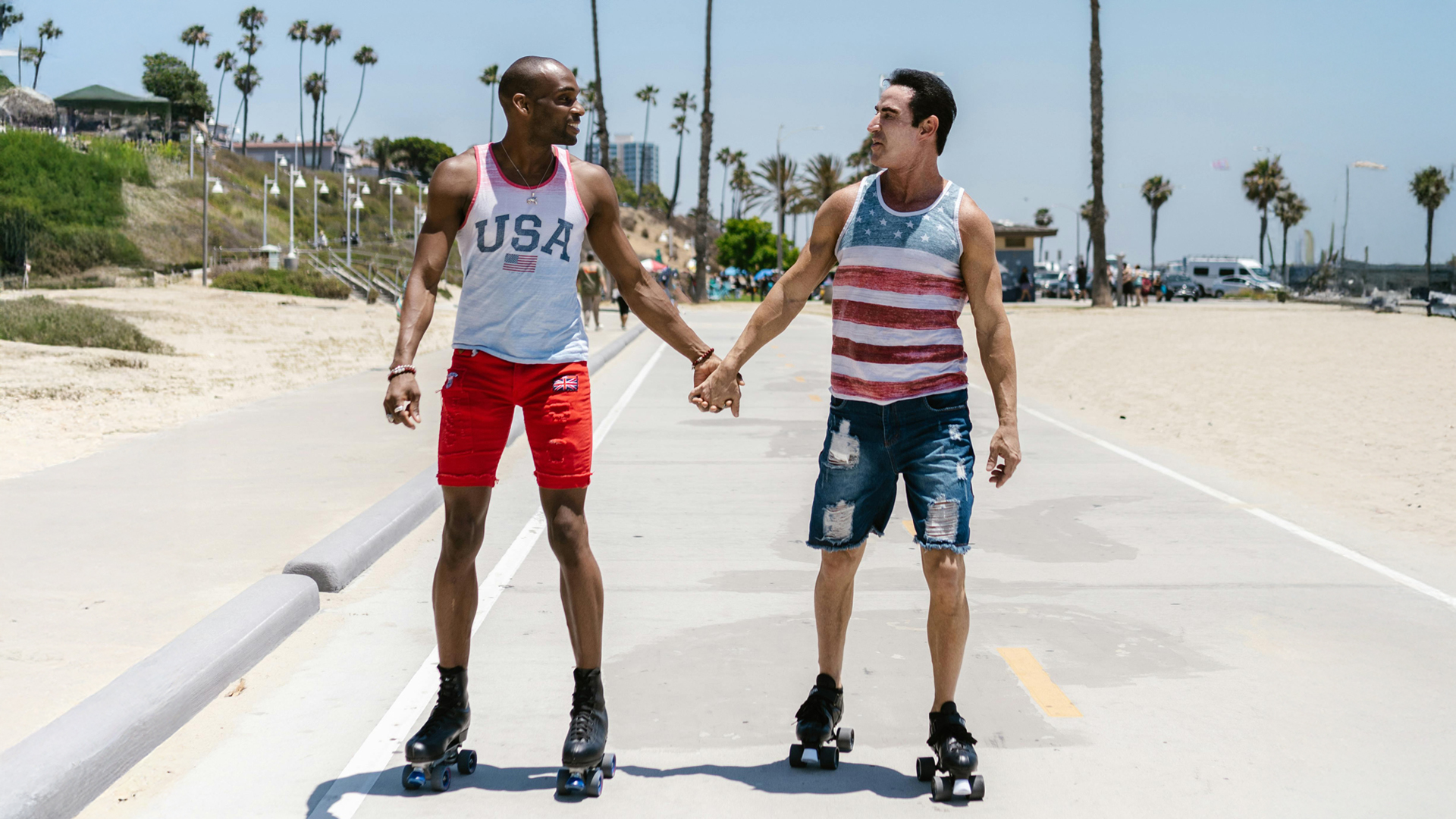 Two men rollerskating along a boardwalk with US-themed tank tops on