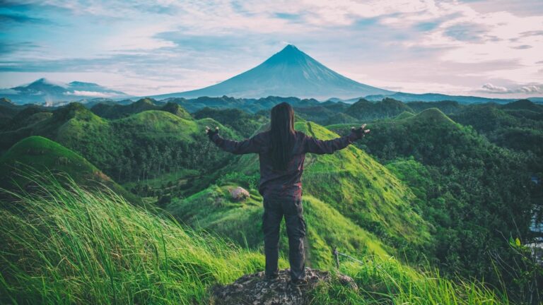 a solo traveller standing before greenery and a volcano with arms outstretched