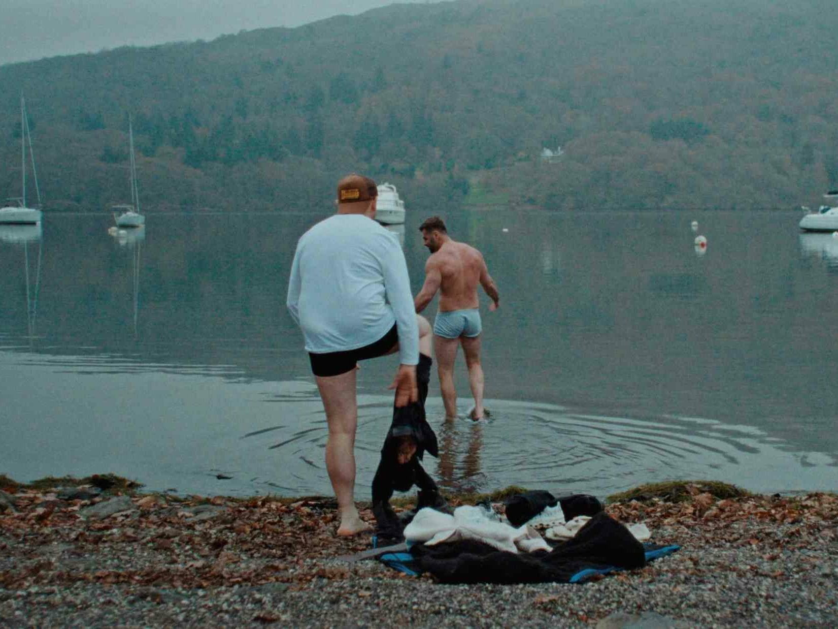 Two men in their underwear stand by a calm lake, one walking towards the water, with boats and misty hills in the background