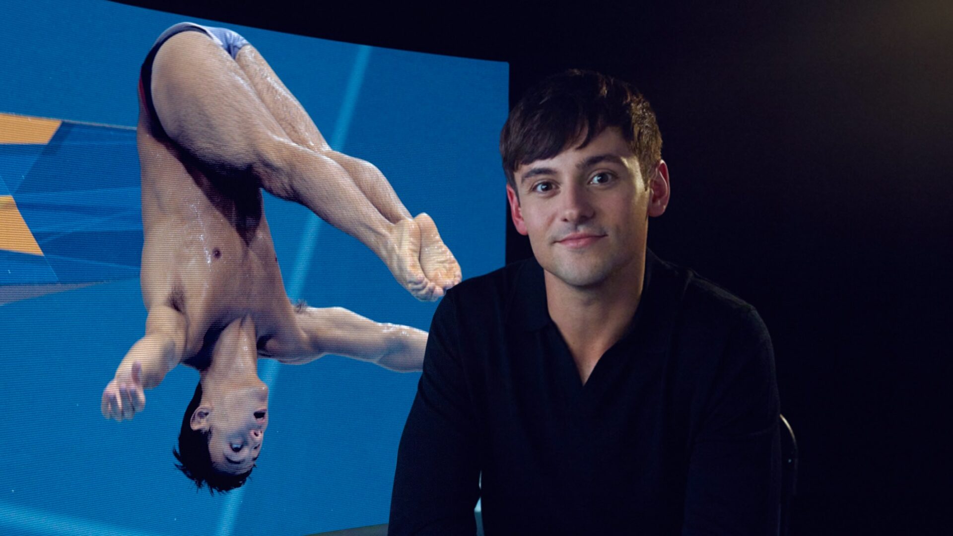 Tom daley in a black shirt sitting before a pic of him diving